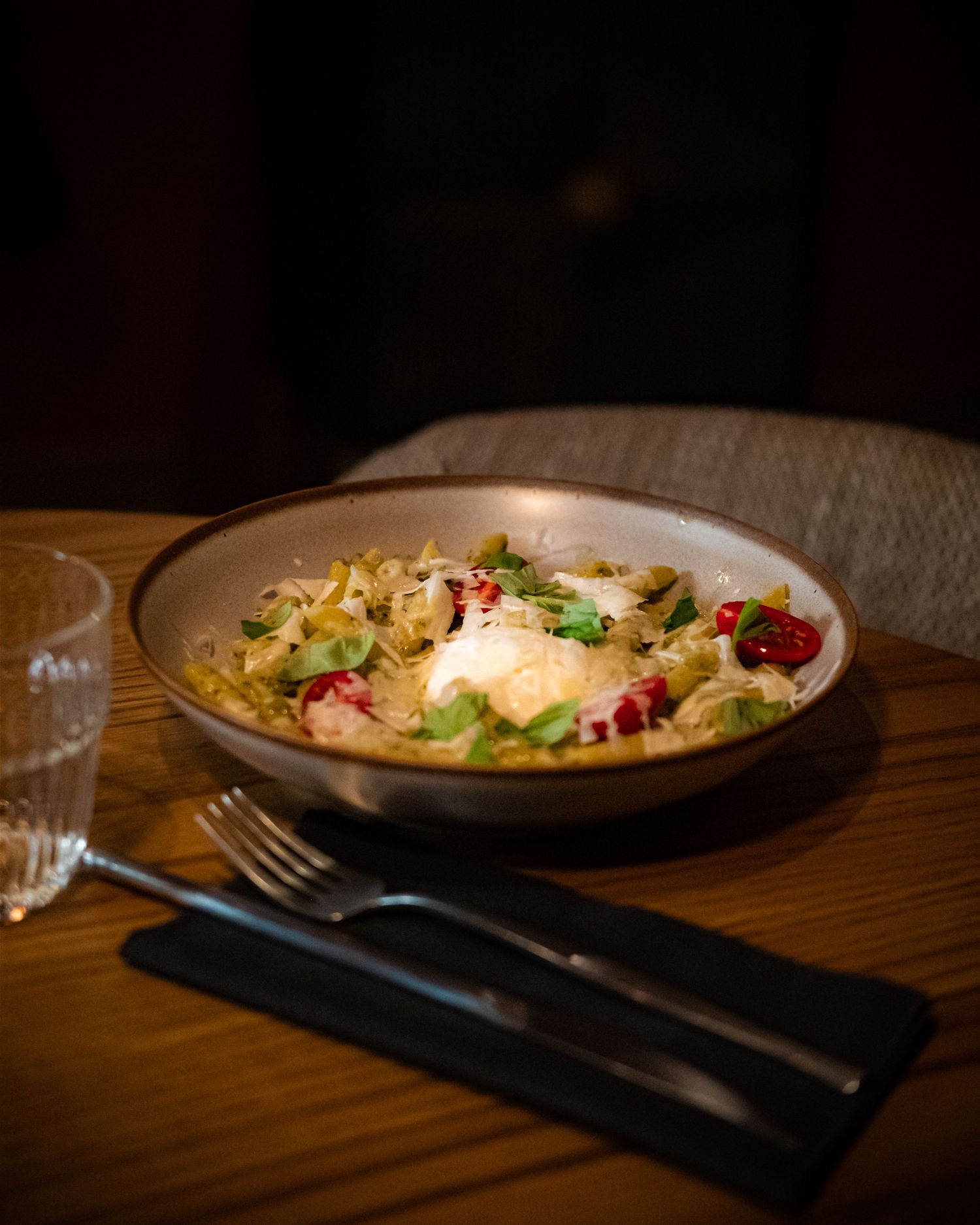 Bowl of pasta with burrata, roasted tomatoes, and fresh herbs on a wooden restaurant table with cutlery.