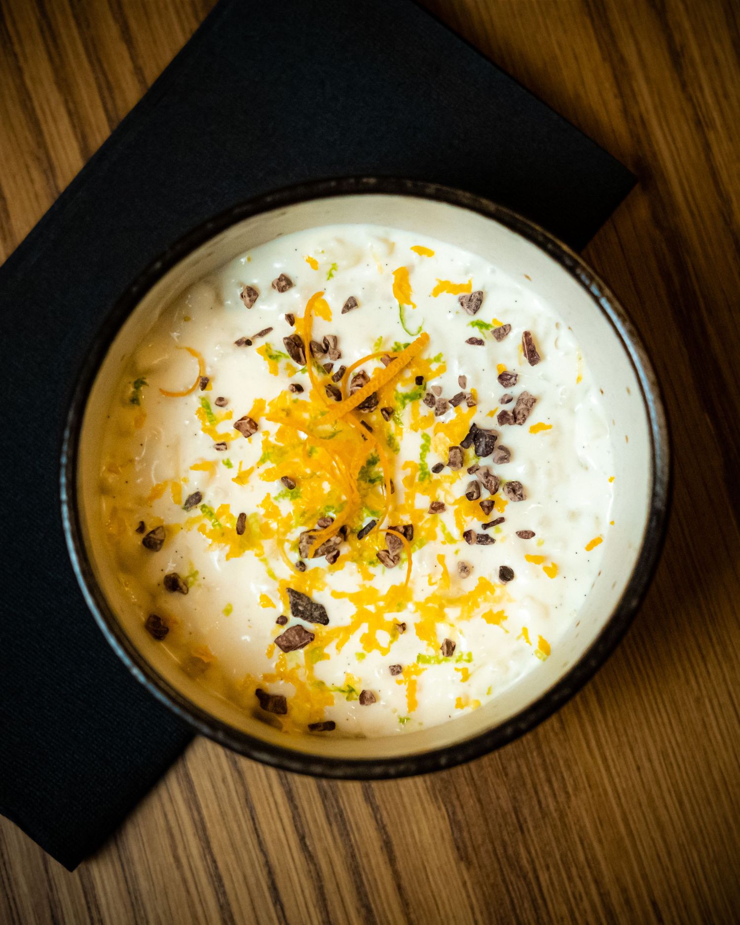 Creamy white dessert bowl topped with orange zest, chopped nuts, and chocolate bits on a wooden table.