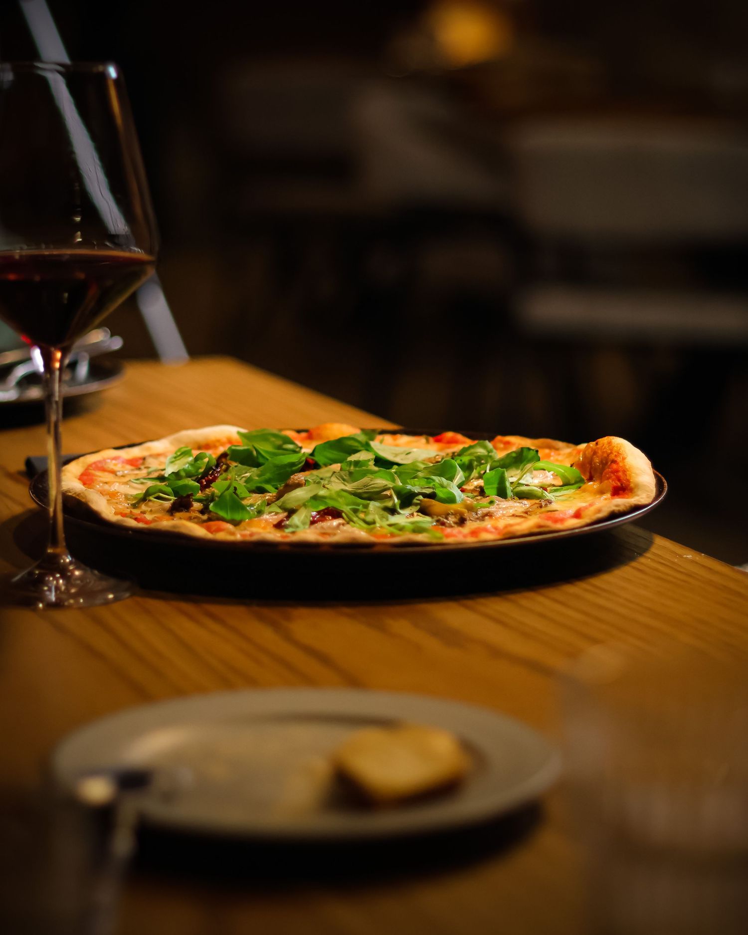 A thin-crust pizza topped with fresh arugula on a wooden table beside a glass of red wine.