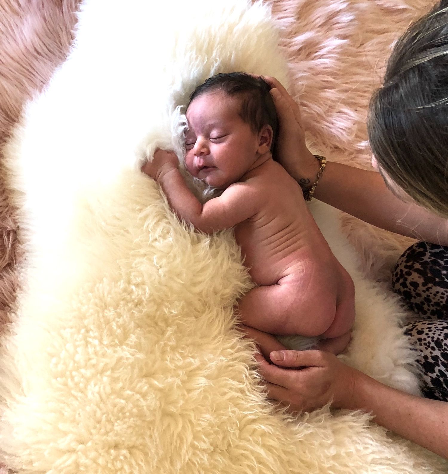 Newborn baby sleeping peacefully on a fluffy cream sheepskin rug, gently held by an adult's hands.