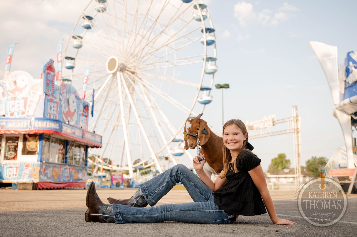 Girl sits with a goat at a county fair, large Ferris wheel and carnival rides visible in the background.