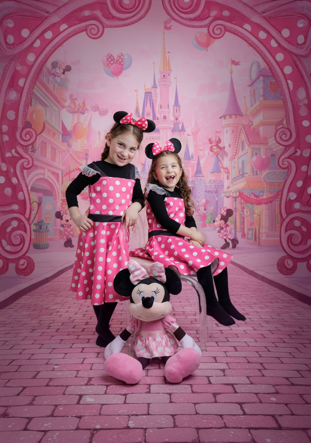 Two girls in matching Minnie Mouse outfits posing with a Minnie plush toy before a pink castle.