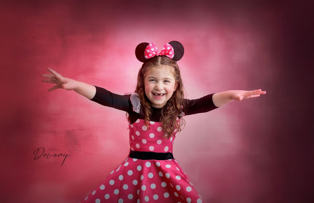 Girl in Minnie Mouse costume with polka dot dress and ears, arms outstretched, smiling on pink background.