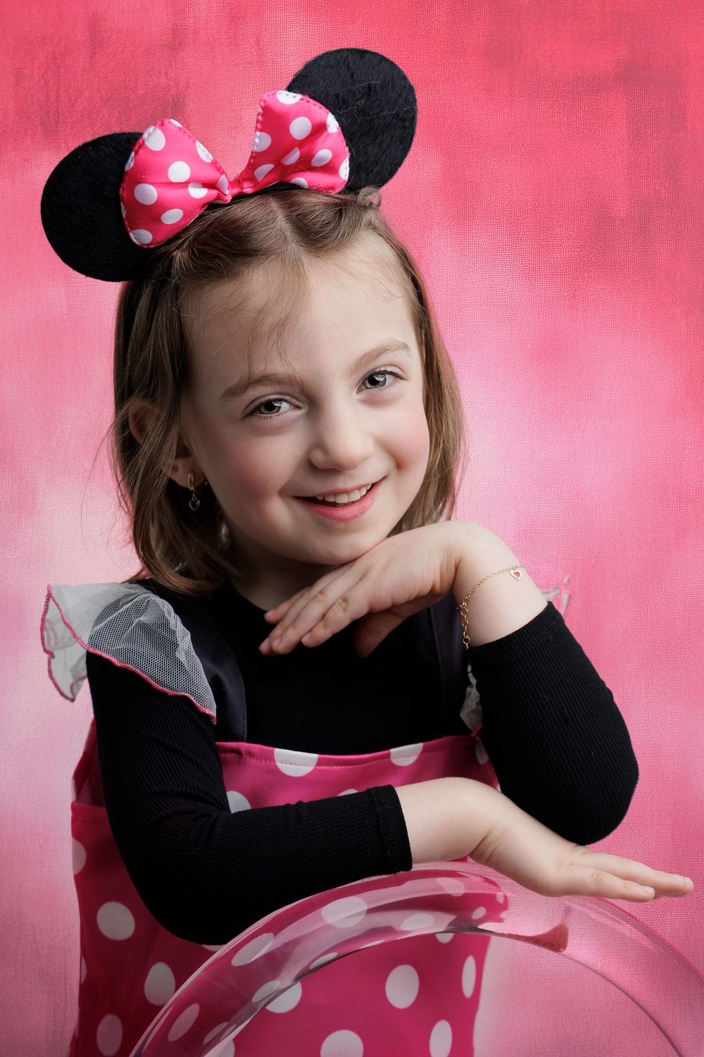 Young girl smiling in Minnie Mouse costume with polka dot ears and pink dress against pink background.