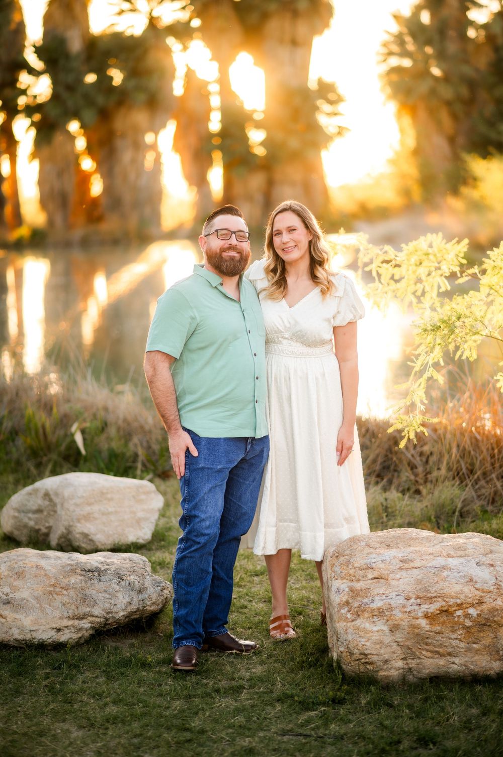 Smiling couple standing together near large rocks with golden sunlight filtering through palm trees.