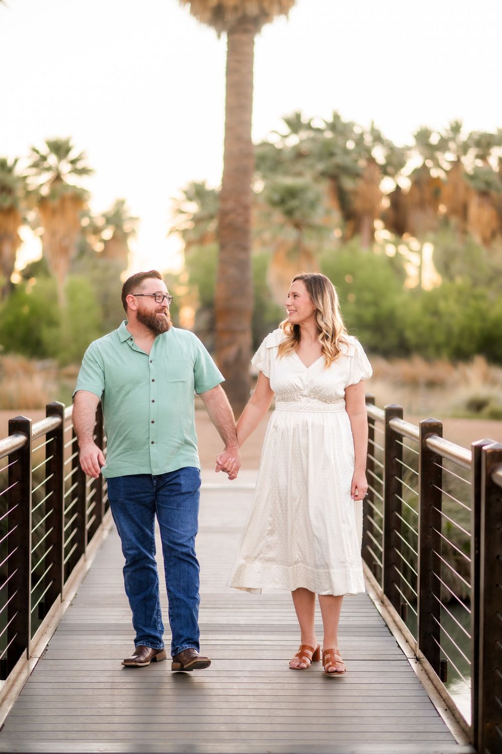 Happy couple holding hands walking on a wooden bridge surrounded by palm trees at golden hour.