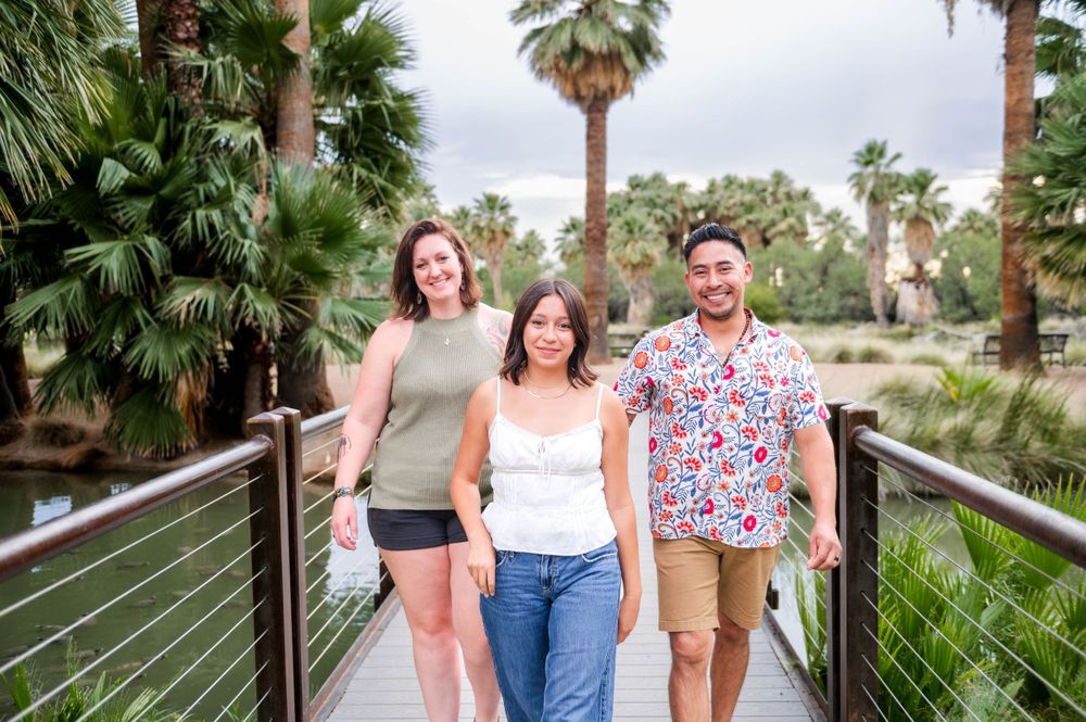 Two women and a man walk together on a bridge surrounded by lush palm trees outdoors.
