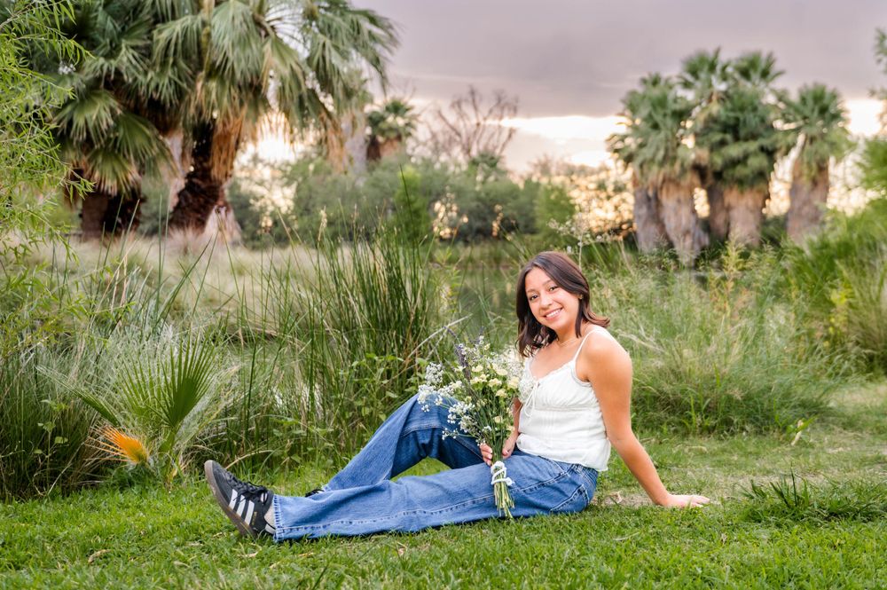 A young woman in a white top and jeans sits on grass holding wildflowers near palm trees.