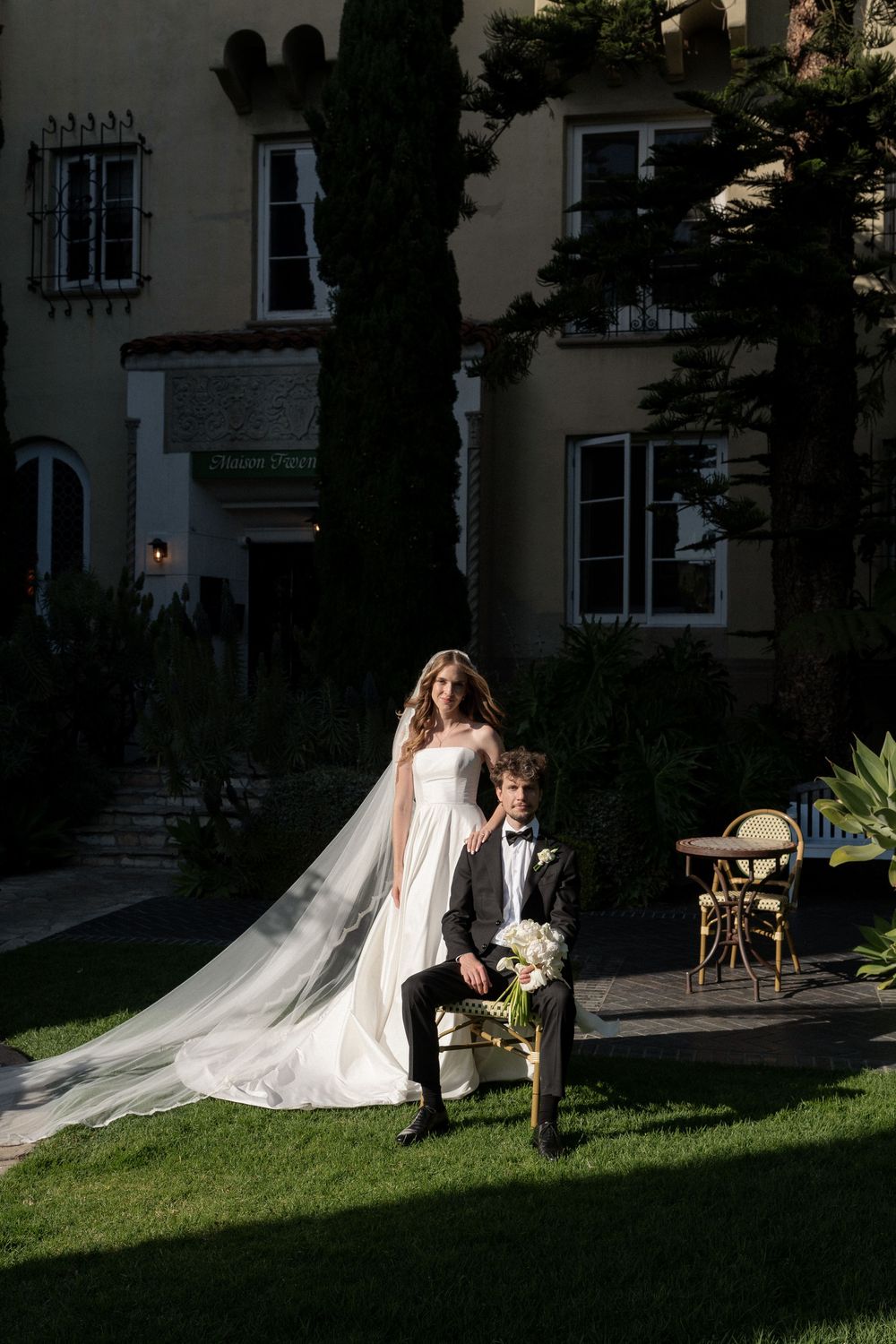 Bride in white gown with long veil stands beside groom in tuxedo on green lawn outside elegant villa.