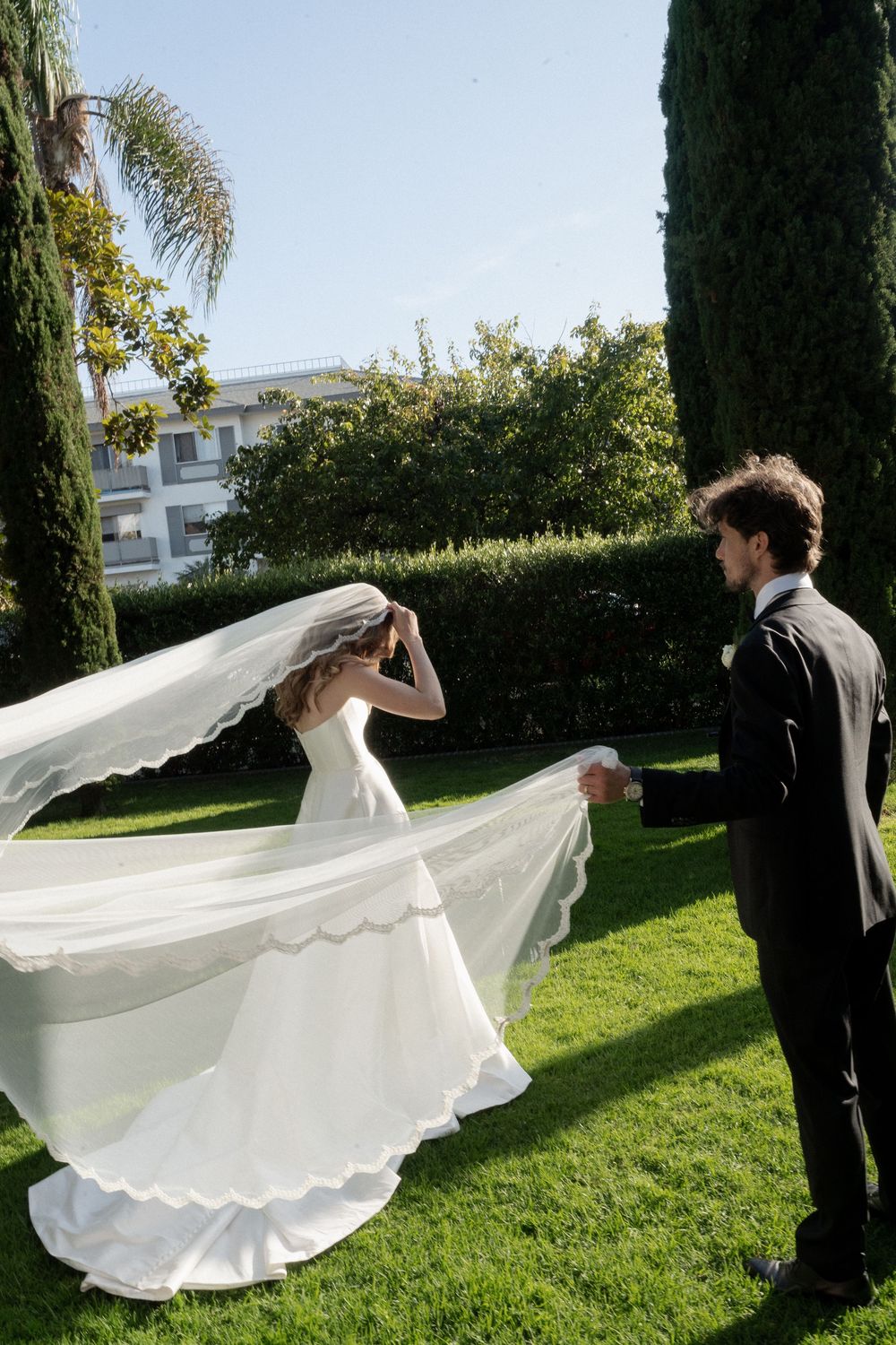 Groom holding bride's flowing veil in a sunlit garden with cypress trees at wedding.