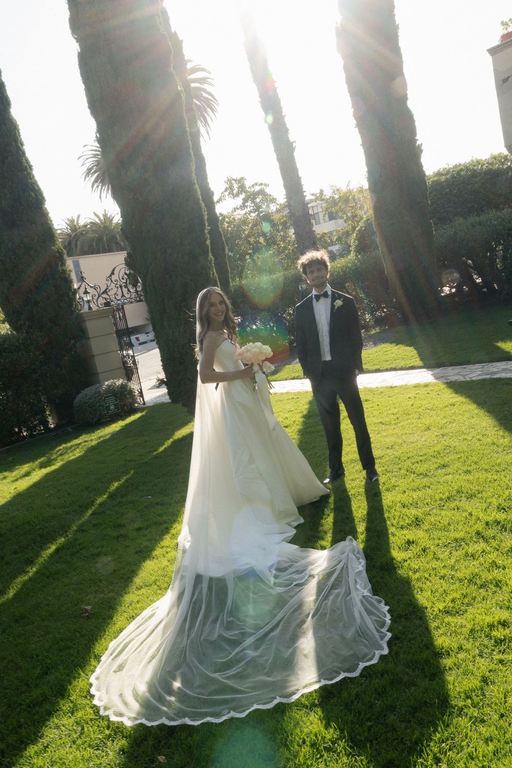 Bride in white gown with long veil and groom in tuxedo stand on green lawn surrounded by tall trees.