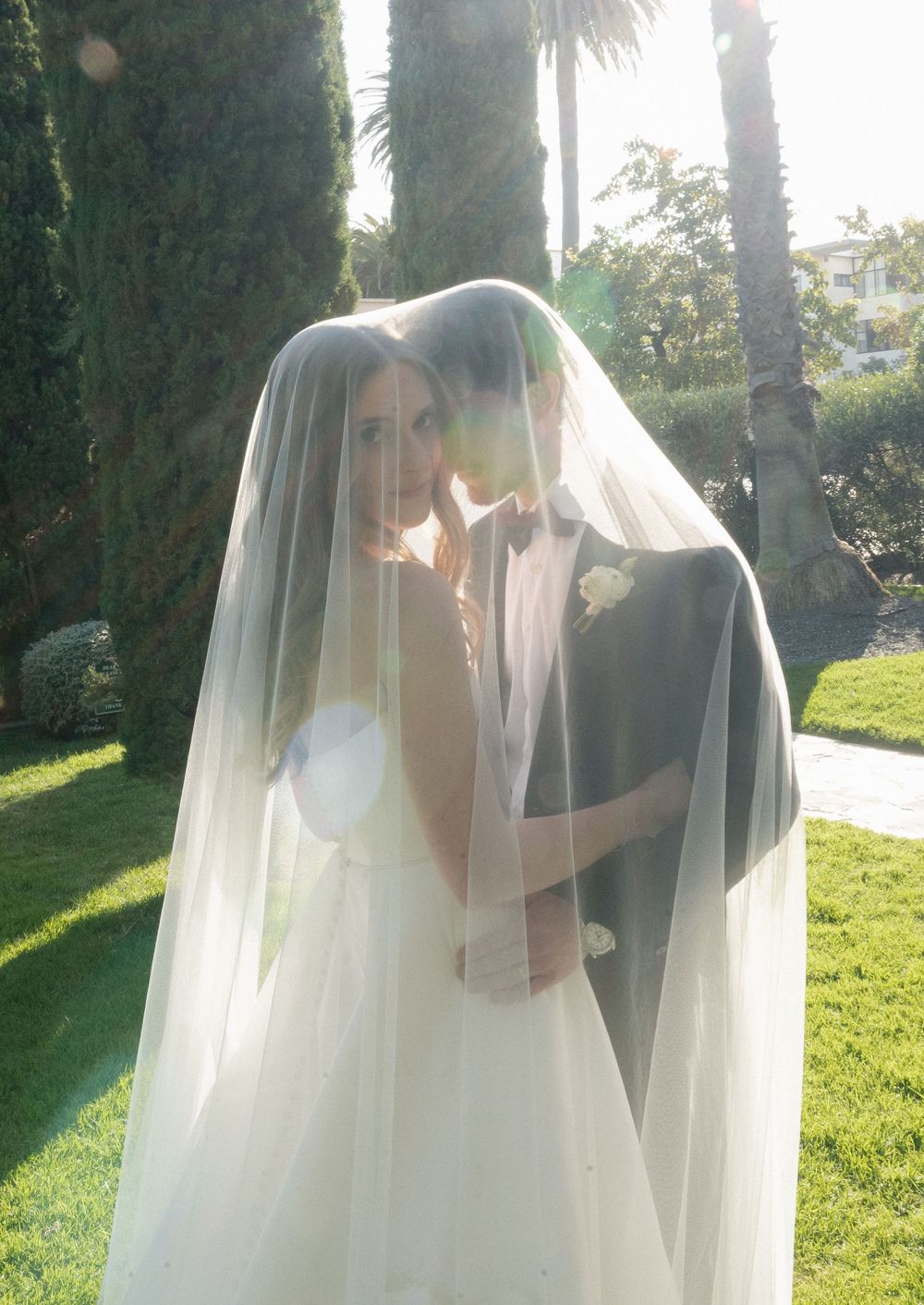 Romantic bride and groom embracing under a sheer wedding veil in golden sunlight outdoors.