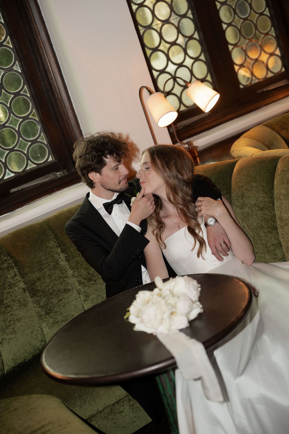 Bride and groom in elegant attire share intimate moment on green velvet sofa with white bouquet nearby.
