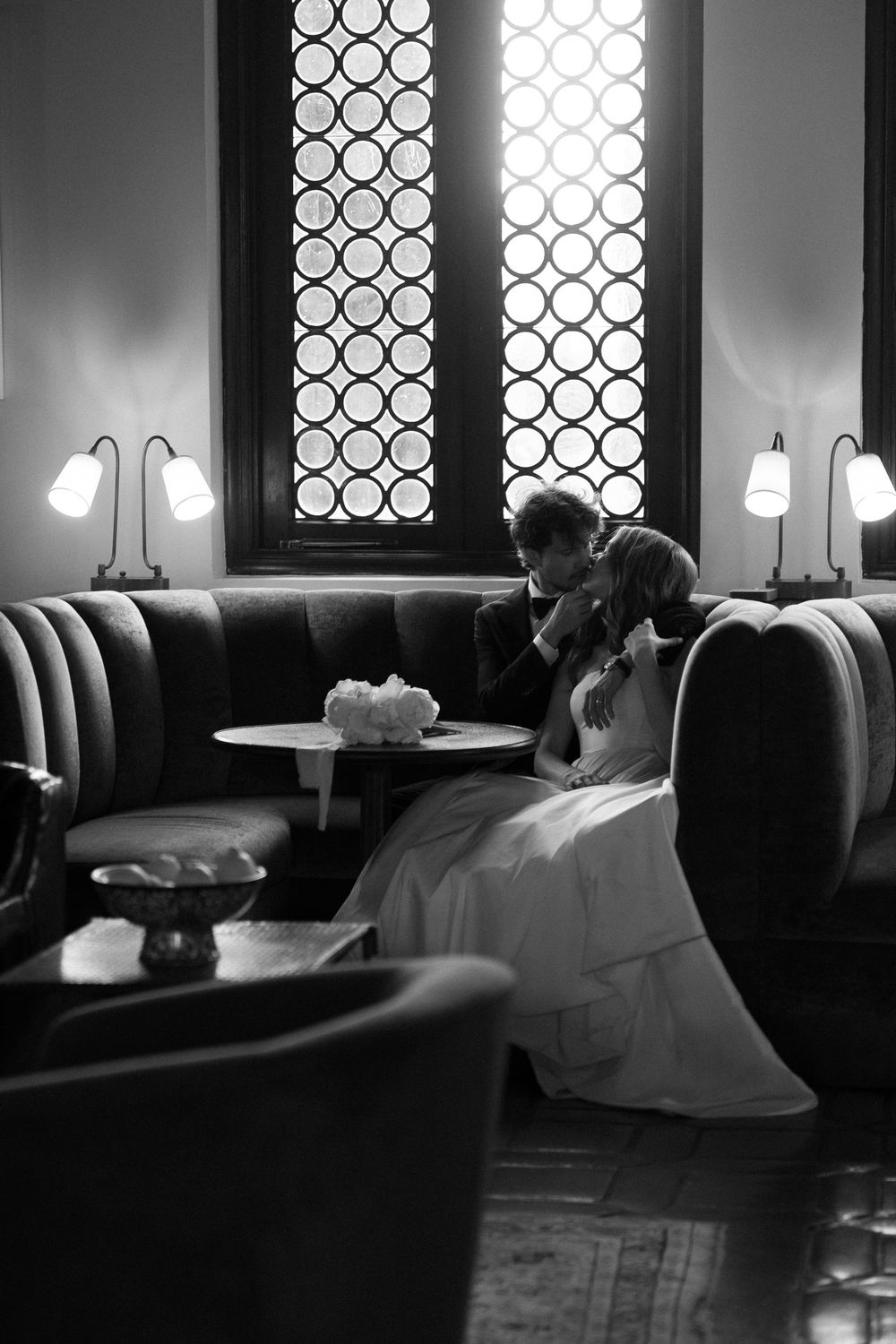 Black and white photo of bride and groom sharing an intimate moment on a velvet sofa in elegant venue.