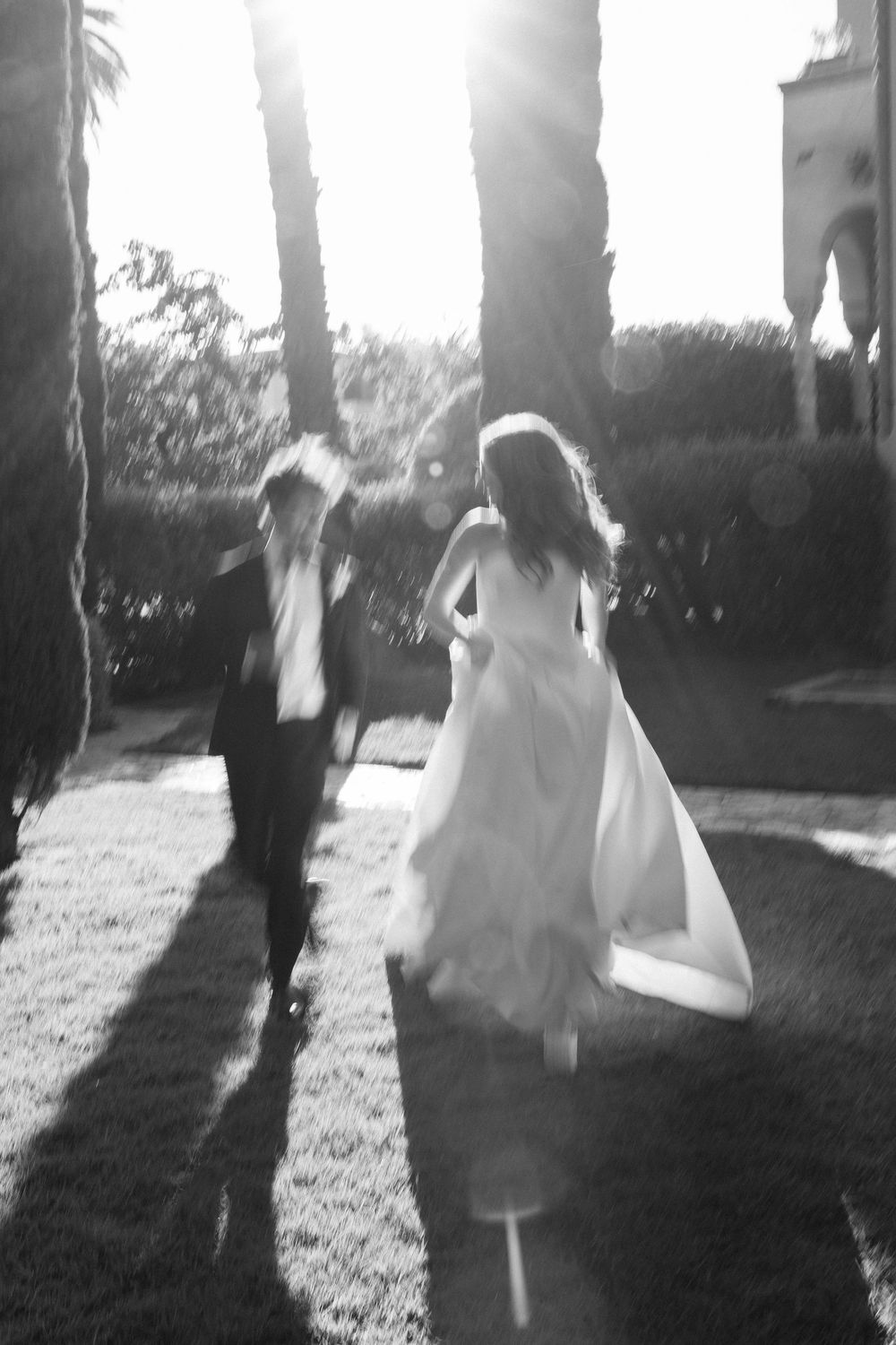 Black and white photo of bride and groom running joyfully through sunlit palm tree-lined grounds.
