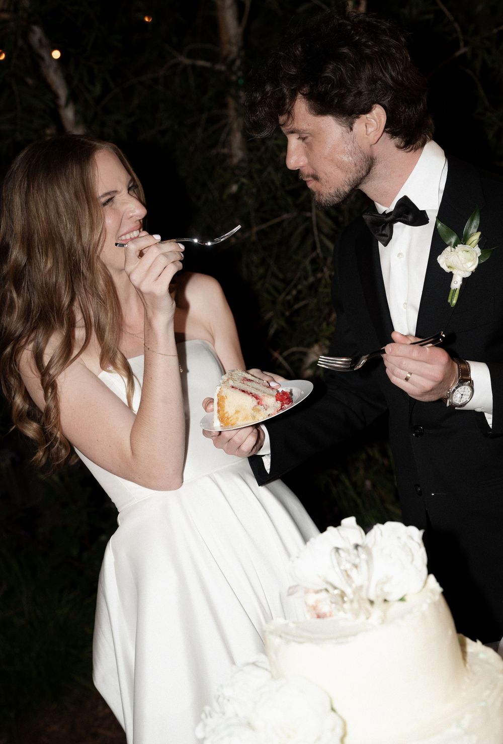 Bride and groom smile while cutting and tasting their white wedding cake at outdoor evening reception.