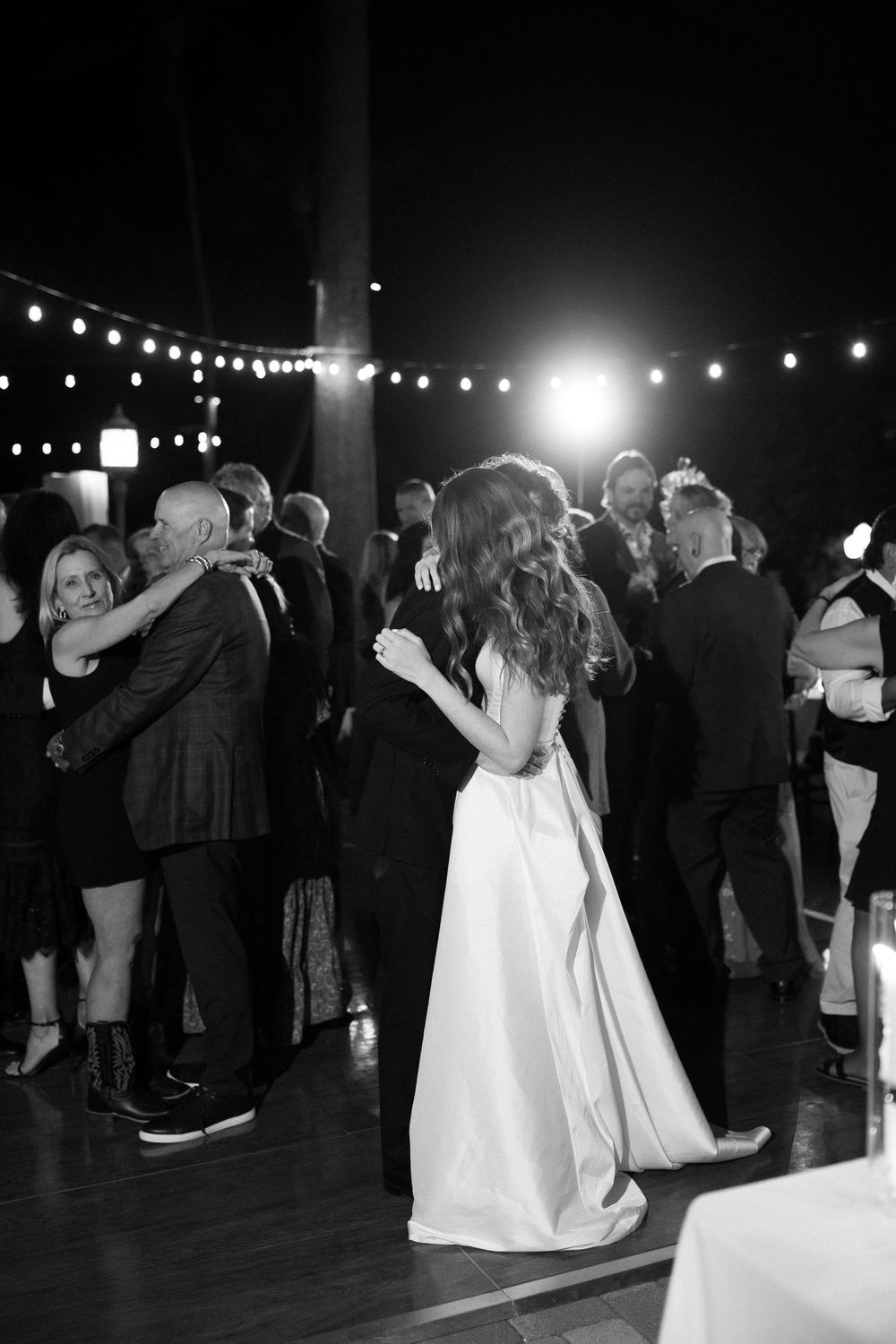 Bride and groom share a dance with guests at a romantic evening wedding reception under string lights.