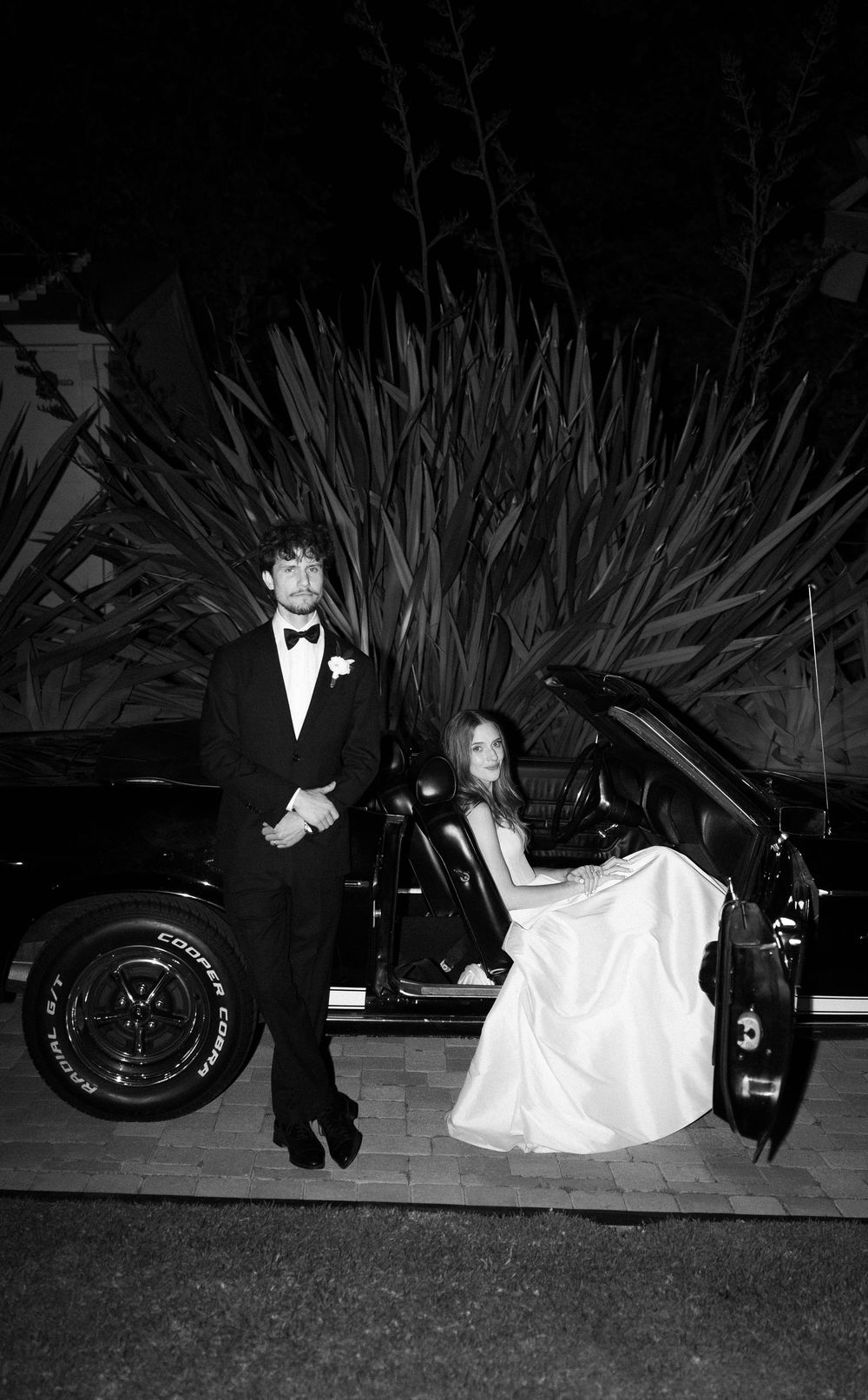 Black and white photo of bride and groom in formal attire posing beside classic car at night.