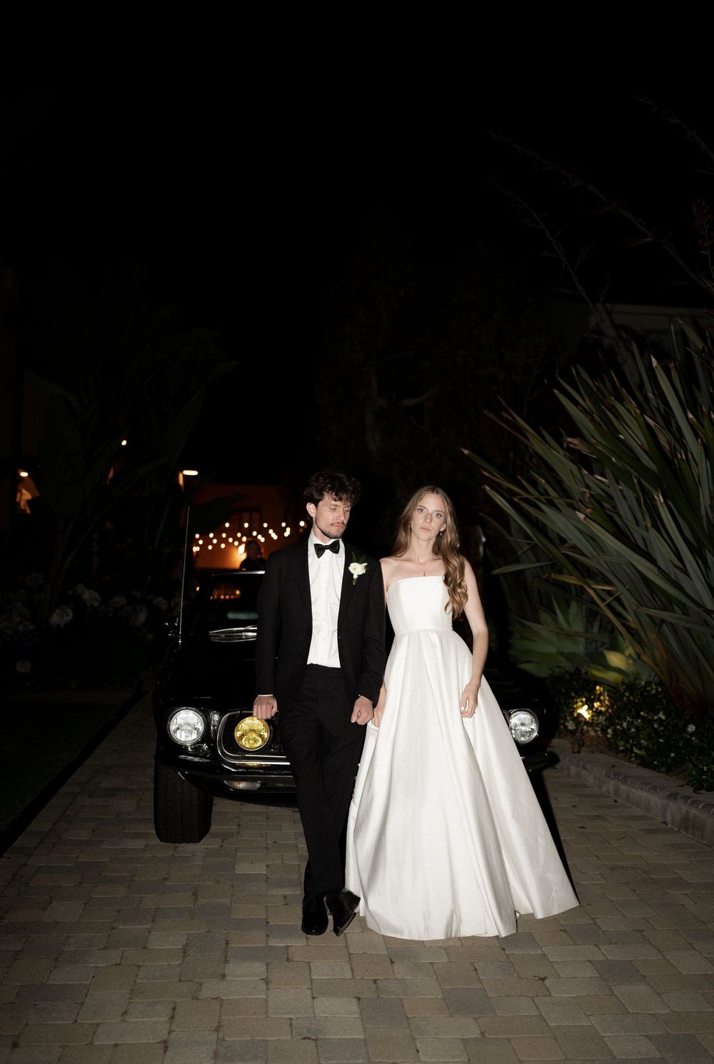 Bride and groom in formal wedding attire posing at night beside a classic black car with string lights.