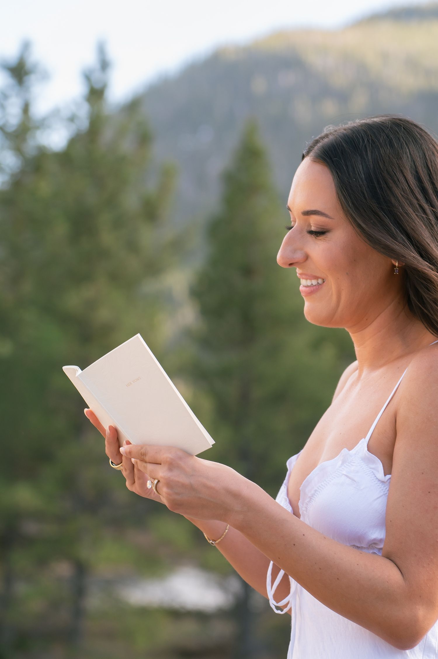 Smiling bride in white dress reads a card outdoors with pine trees and mountains in background.