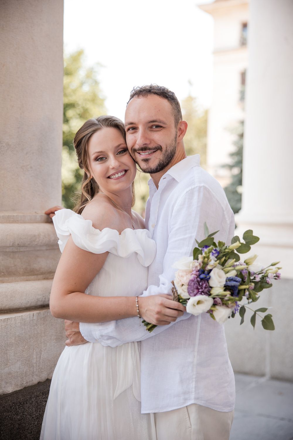 Smiling bride and groom in white attire embracing outdoors, holding a bouquet of purple and white flowers near stone columns.