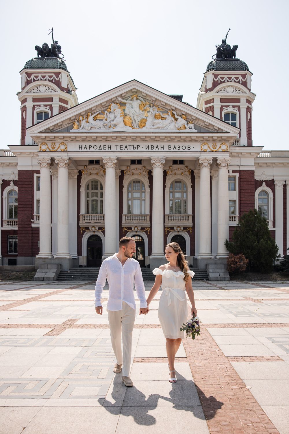Wedding couple in white attire holding hands before Ivan Vazov National Theatre in Sofia, Bulgaria, on a sunny day.