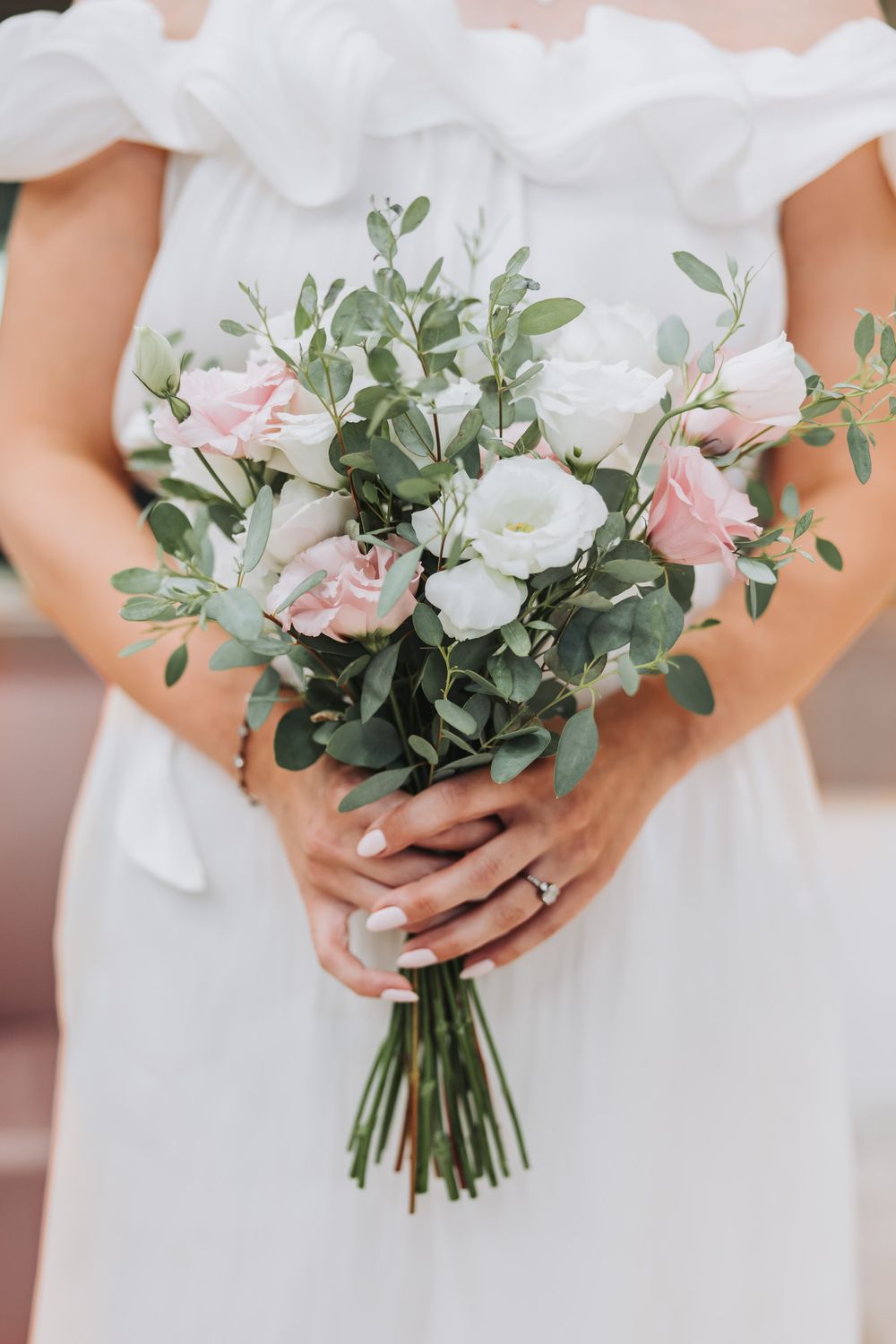 Bride in white off-shoulder dress holding elegant bouquet of pink and white roses with eucalyptus greenery.