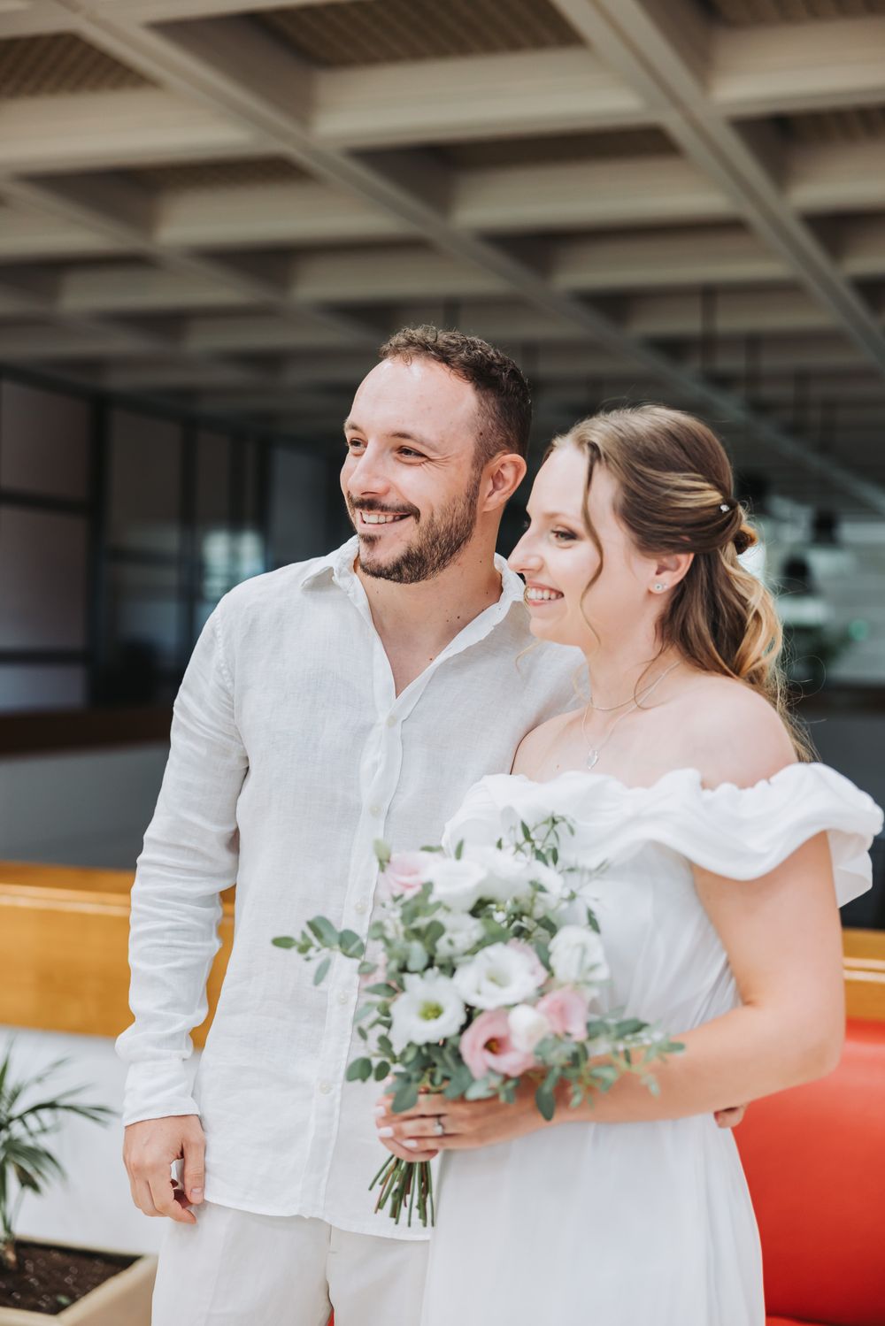Happy couple in white wedding attire smiling outdoors, bride holding a bouquet of pink and white flowers.
