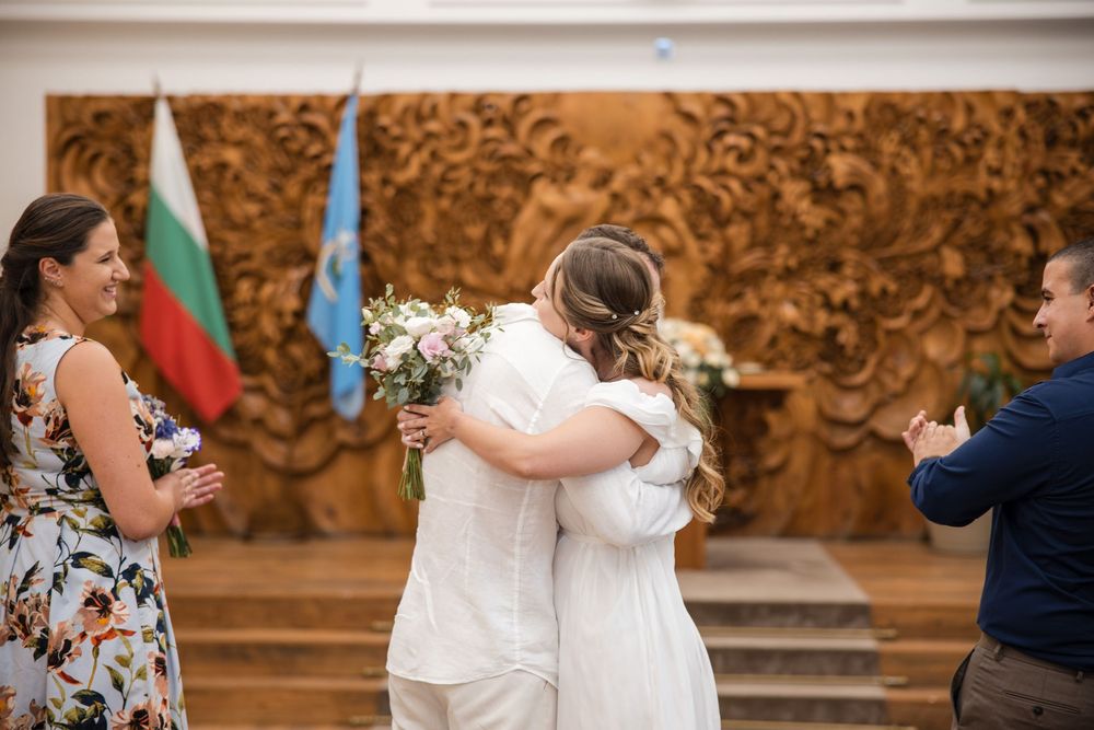 Bride and groom in white embrace during a wedding ceremony in a hall decorated with a Bulgarian flag and wooden wall art.