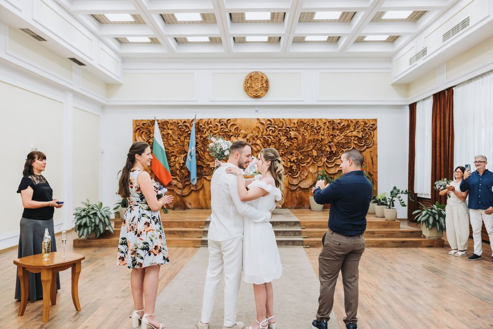 Couple sharing first kiss at civil wedding ceremony in elegant hall with Bulgarian flag and wooden decor, surrounded by guests.