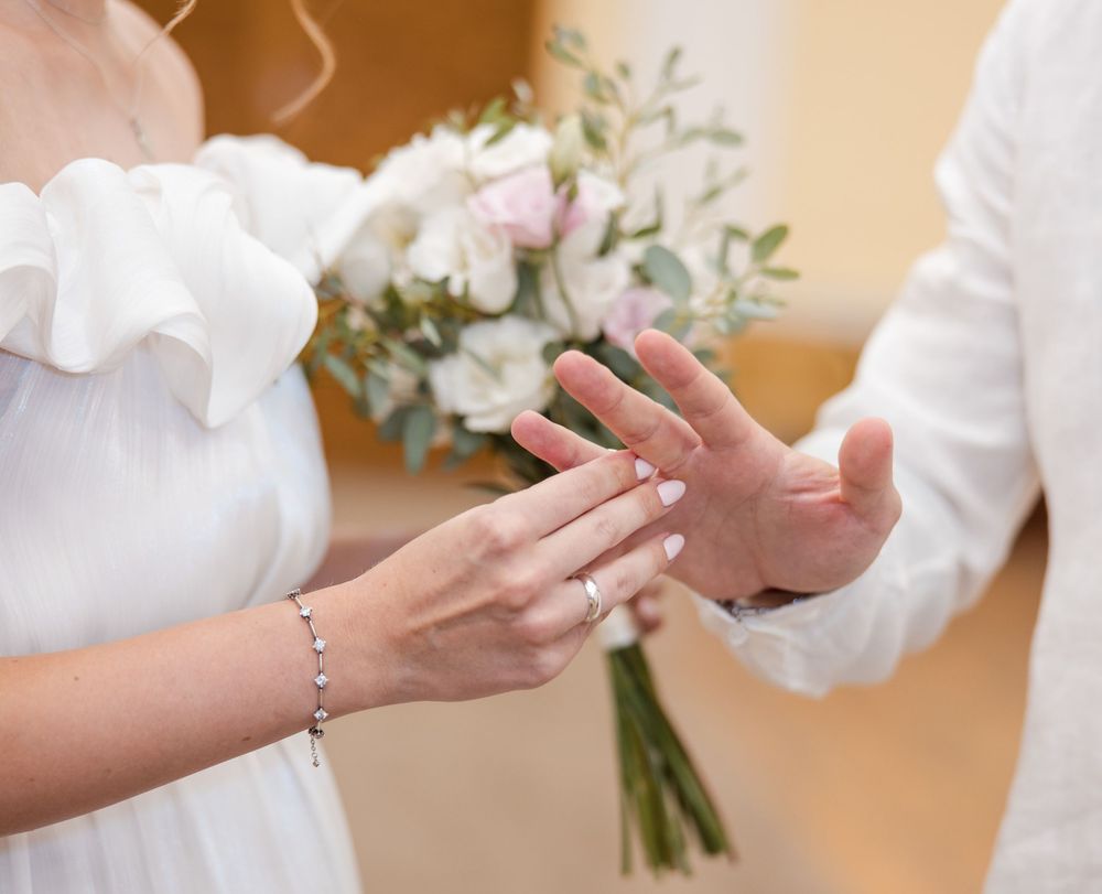 Bride placing wedding ring on groom's finger during ceremony, holding white floral bouquet with pink roses.