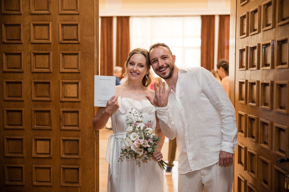 Newlyweds in white attire smile and show wedding rings and wedding certificate while holding bouquet by wooden doors.
