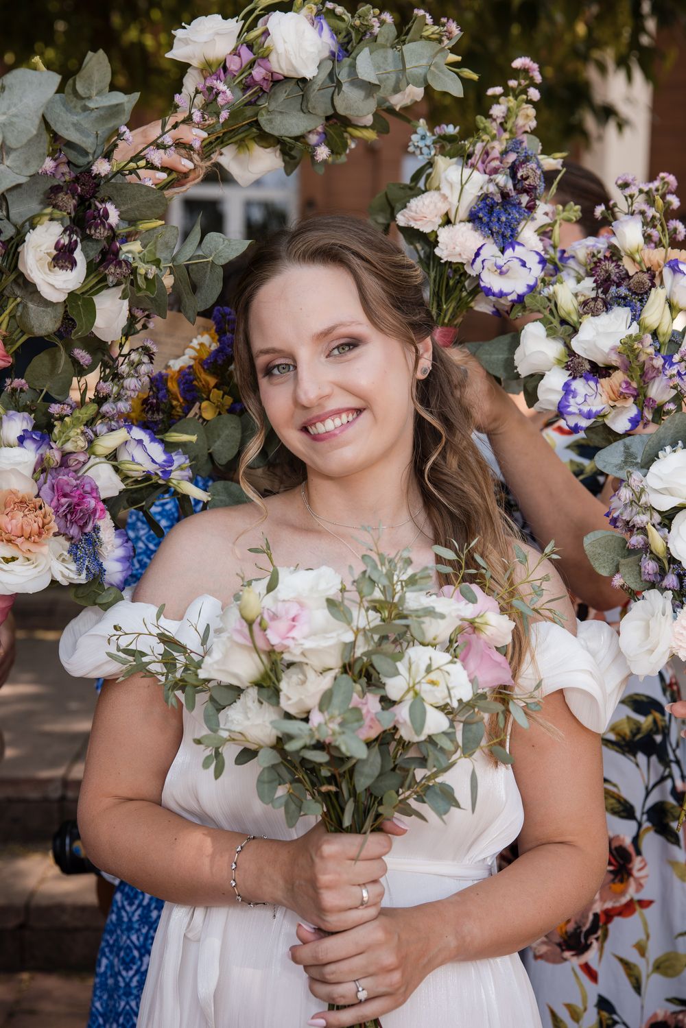 Smiling bride in white off-shoulder dress holds bouquet of white and pink roses, standing before colorful floral arch.