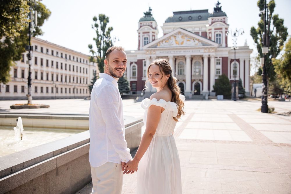 Couple in white attire holding hands and smiling in front of a grand historic building on a sunny day.