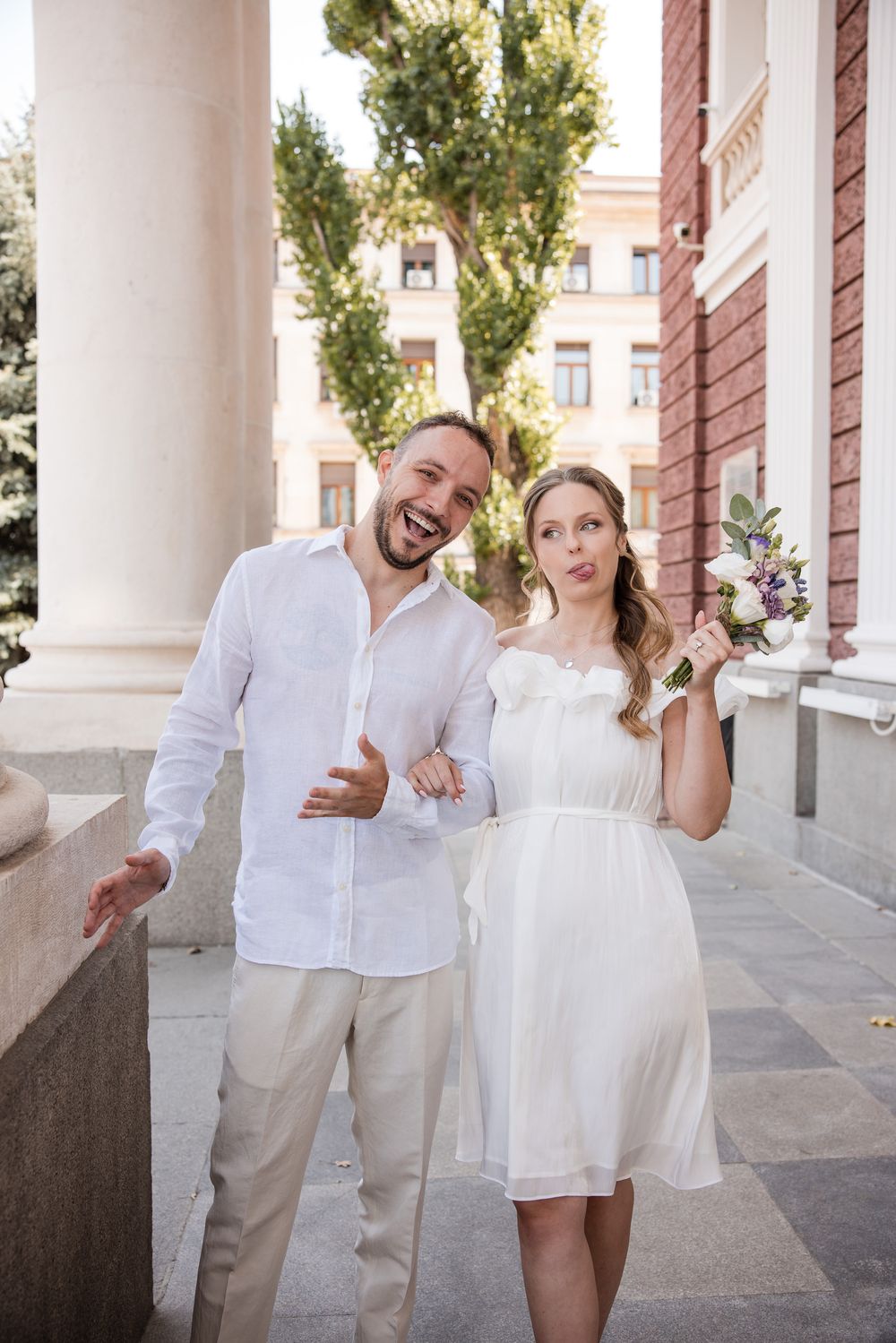 Happy couple in white attire posing outdoors, bride holding bouquet, groom gesturing, near classical columns.