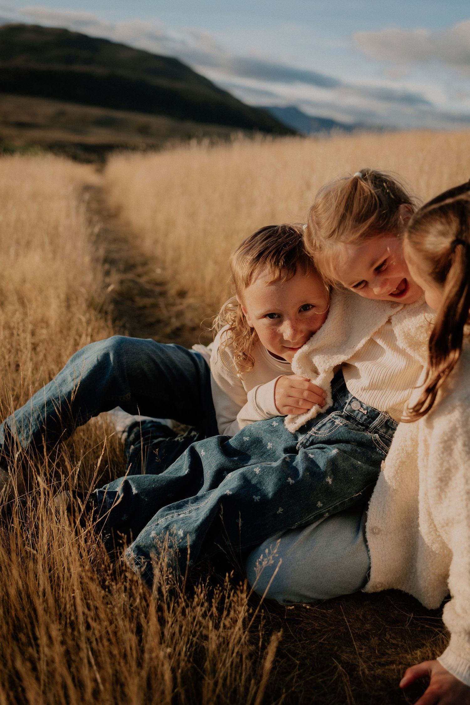 Three children playing and laughing together in a golden grass field with mountains in the background.