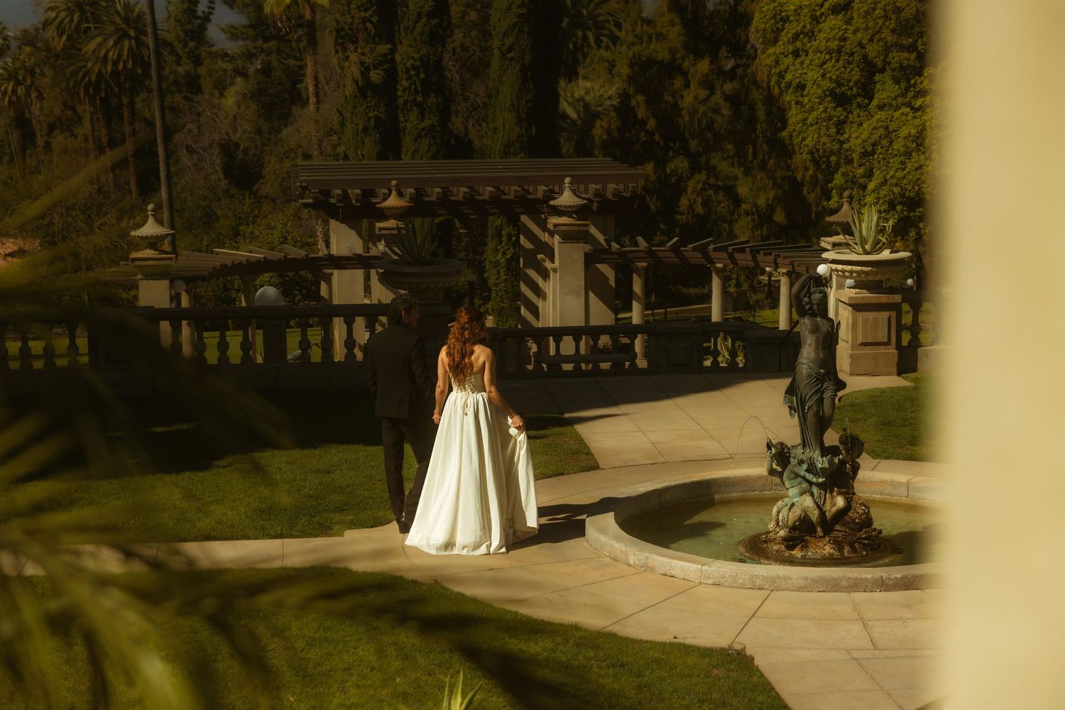 A bride and groom walk hand in hand through the Kimberly Crest House wedding venue with a fountain and lush greenery.