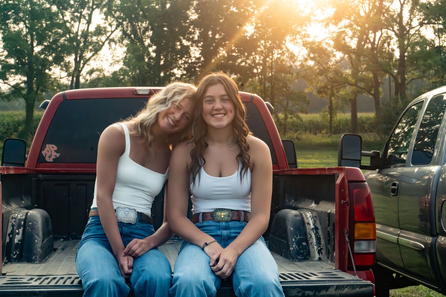 Two young women in white tops and jeans sitting together on a red pickup truck tailgate at golden hour.