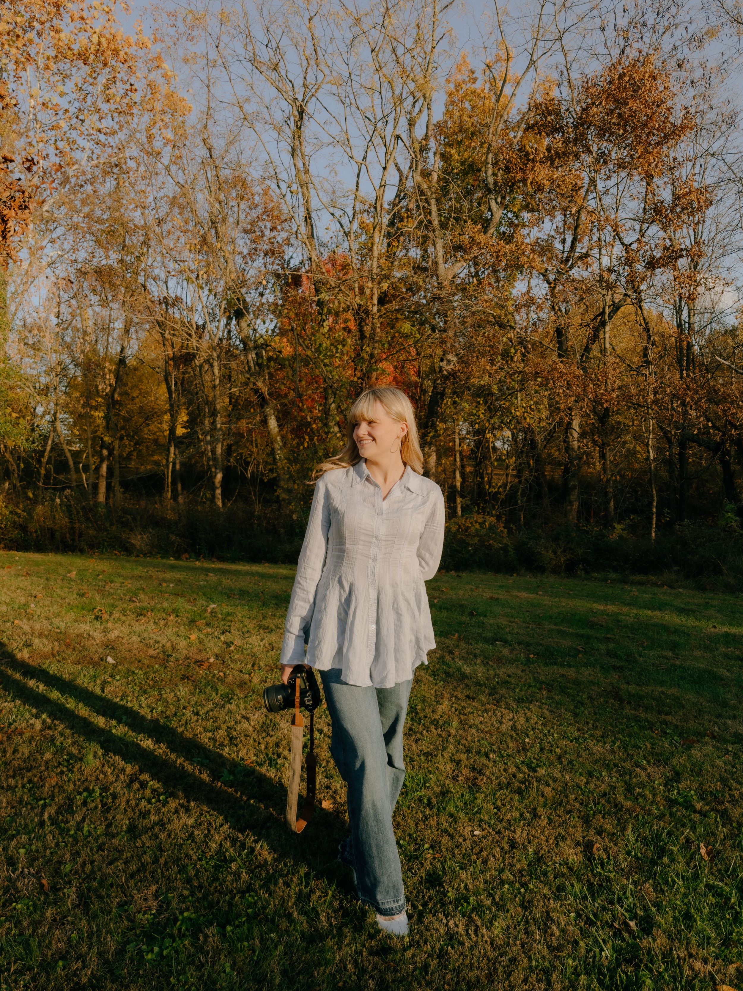 Woman in white blouse and jeans holding camera walks through lush autumn park with golden foliage.