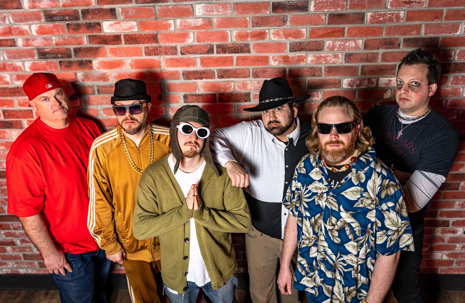 Seven musicians in stylish hats and sunglasses pose against a brick wall for a band photo.