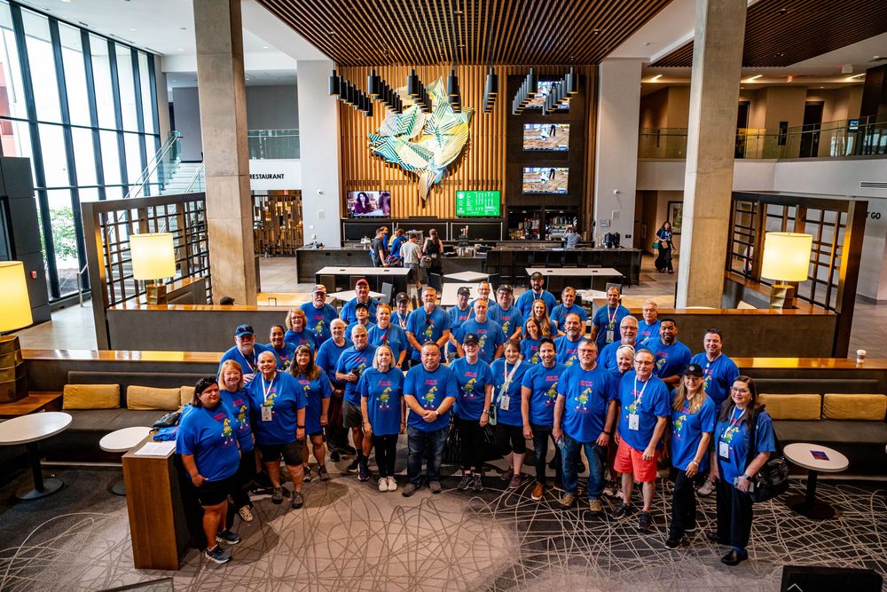 Large group of volunteers in matching blue t-shirts pose together in modern hotel lobby atrium.