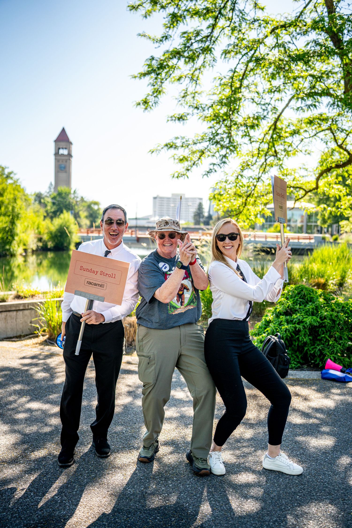 Three people posing playfully outdoors near a river with a clock tower visible in the background.