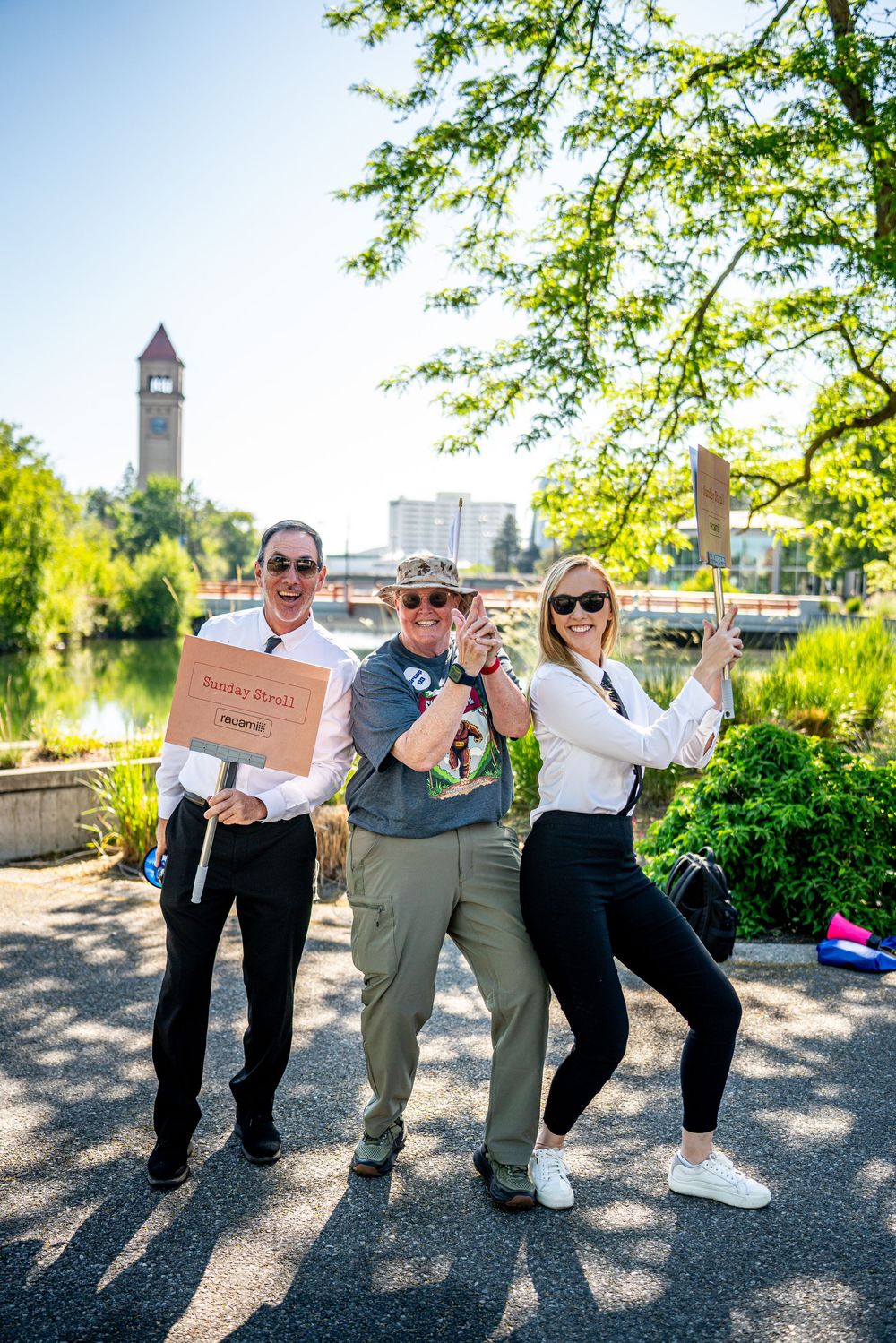 Three people posing playfully outdoors near a river with a clock tower visible in the background.