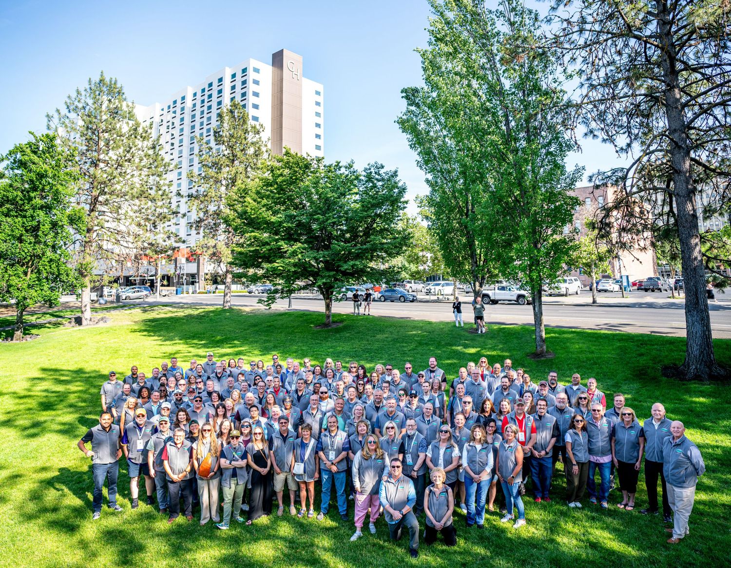 Large group of employees in matching gray shirts gathered outdoors on a sunny green lawn.