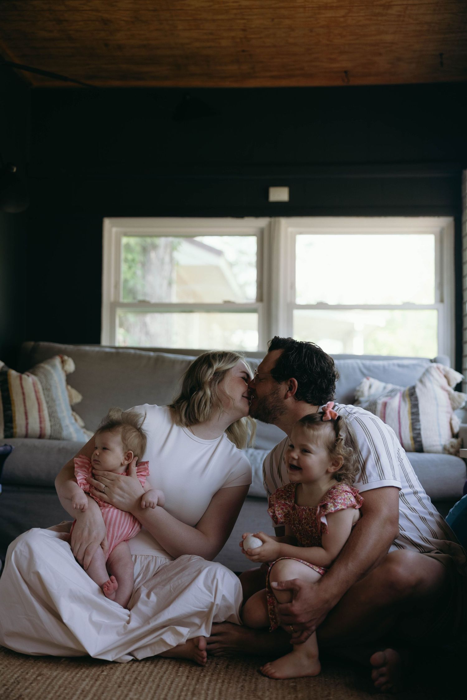 A loving family sits on the floor, parents kissing while holding their two young daughters in a cozy living room.