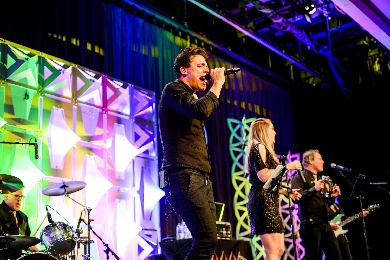 Energetic band performing on a colorful illuminated stage with singers and guitarist entertaining a crowd.
