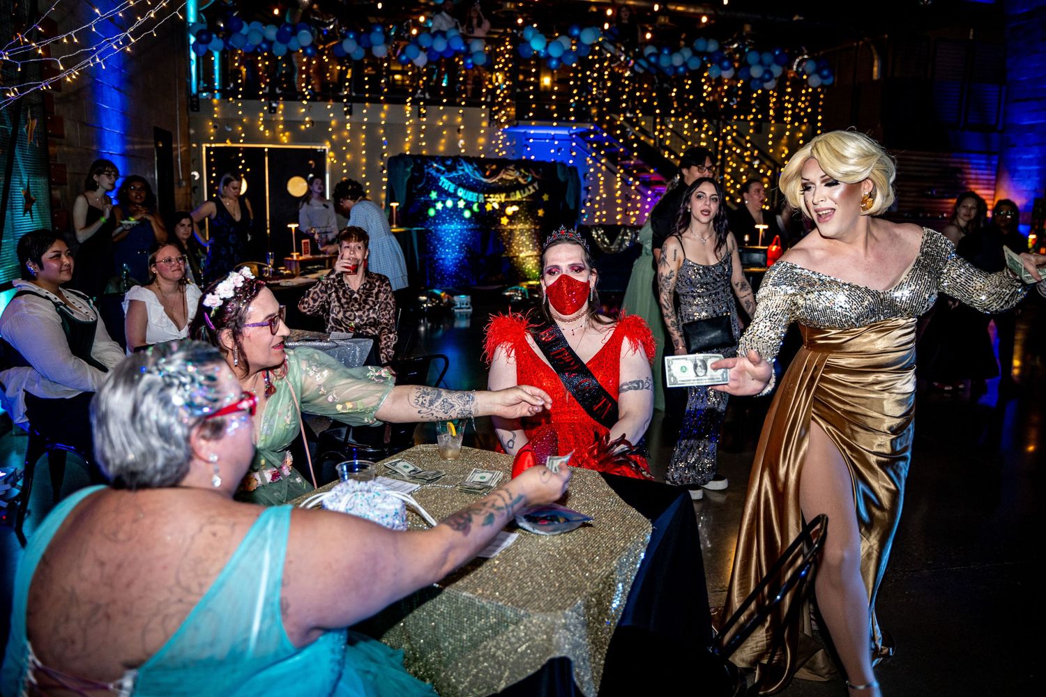 Drag performers interact with guests at glamorous evening gala event with twinkling fairy lights backdrop.