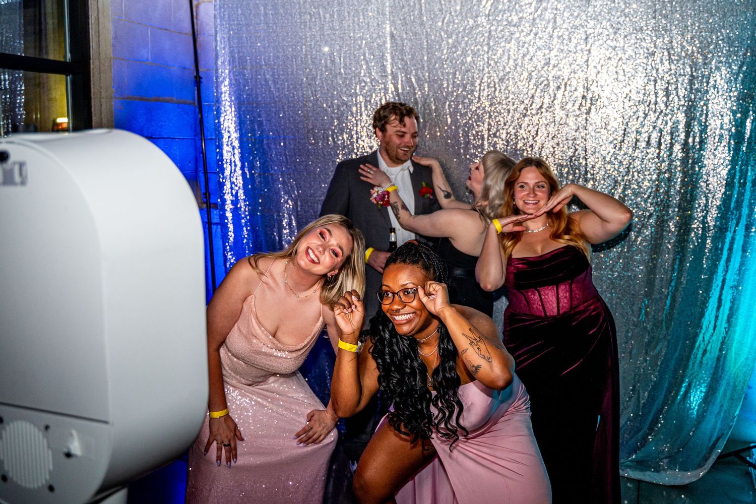 Four friends pose and laugh together in front of silver sequin photo booth backdrop at formal event.