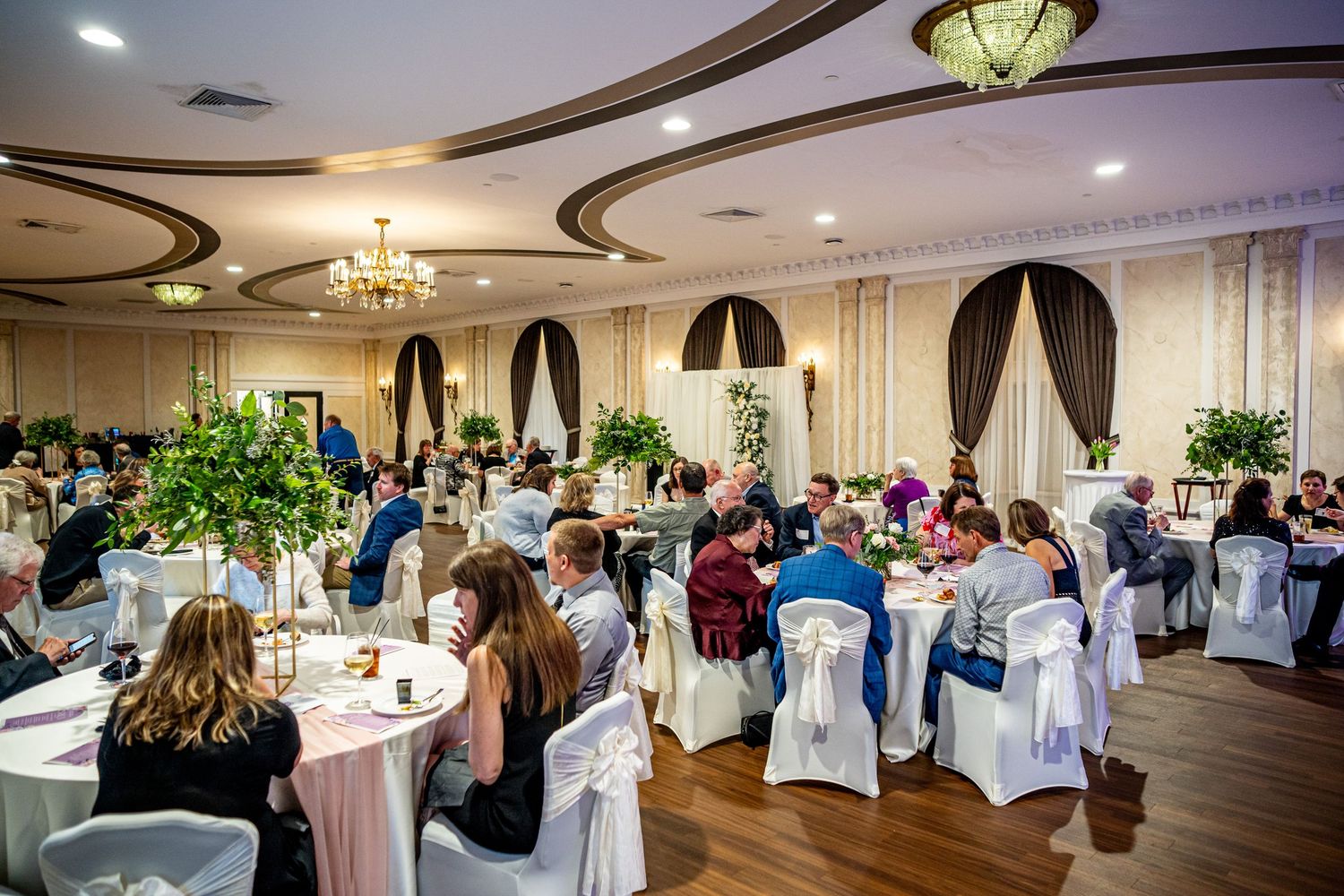 Elegant banquet hall filled with guests dining at white linen tables with floral centerpieces during formal event.