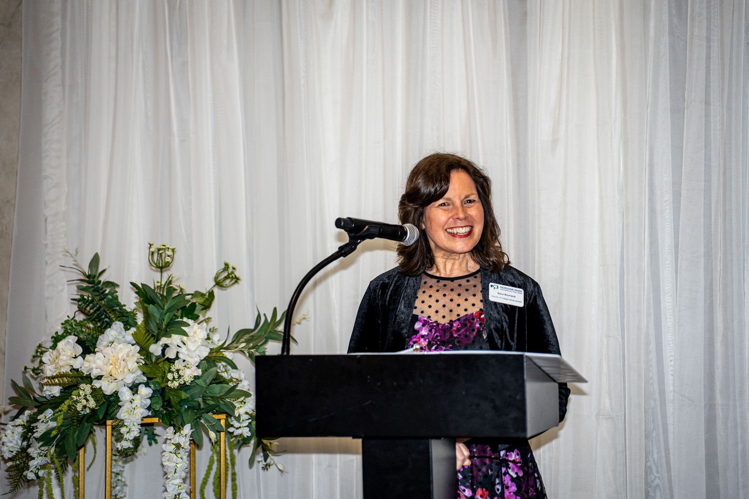 Woman in floral dress smiles while speaking at podium with white floral arrangements at formal banquet.